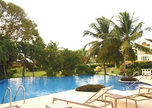 Una piscina con sillones de descanso, palmeras y un edificio al fondo.