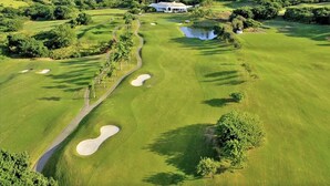 Golfkenttä, jossa on hiekkabunkkereita, lampi ja kerhotalo, ja sen ympärillä on vehreää kasvillisuutta sekä asuinrakennuksia.