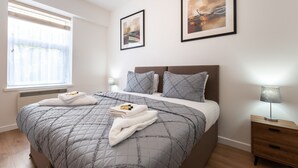 Una cama bien hecha con colcha gris, dos almohadas y toallas dobladas. Hay una mesita de noche con una lámpara, una ventana con persianas, y dos cuadros en la pared.