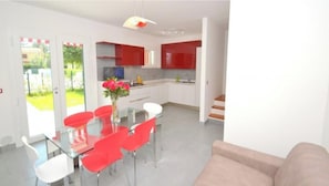 A modern kitchen with red cabinets, a glass dining table with red chairs, and a beige sofa in a bright living area.