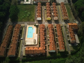An aerial view of a residential area with multiple rows of houses, a central swimming pool, and a road running through the middle.
