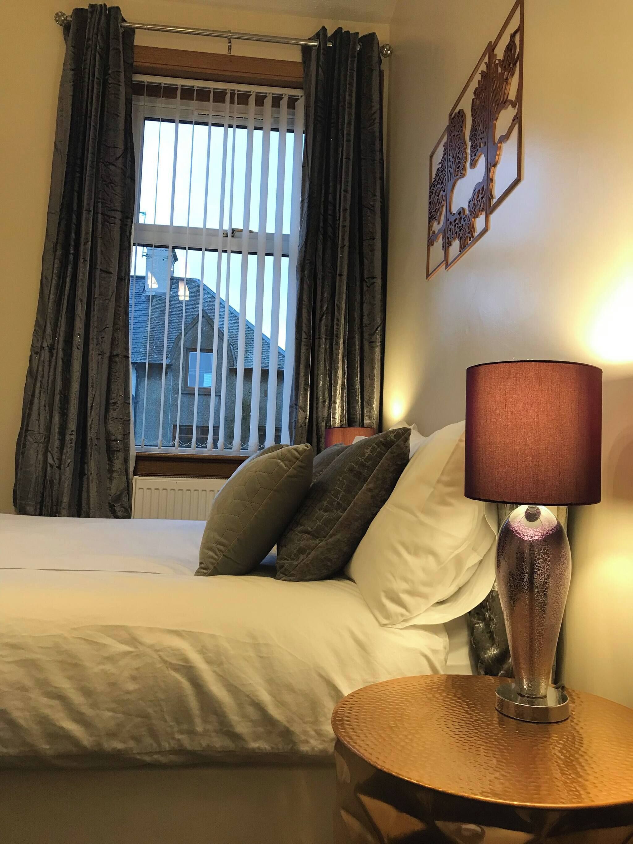 Quarto de hotel com cama, uma mesa redonda com abajur e uma janela com cortinas.