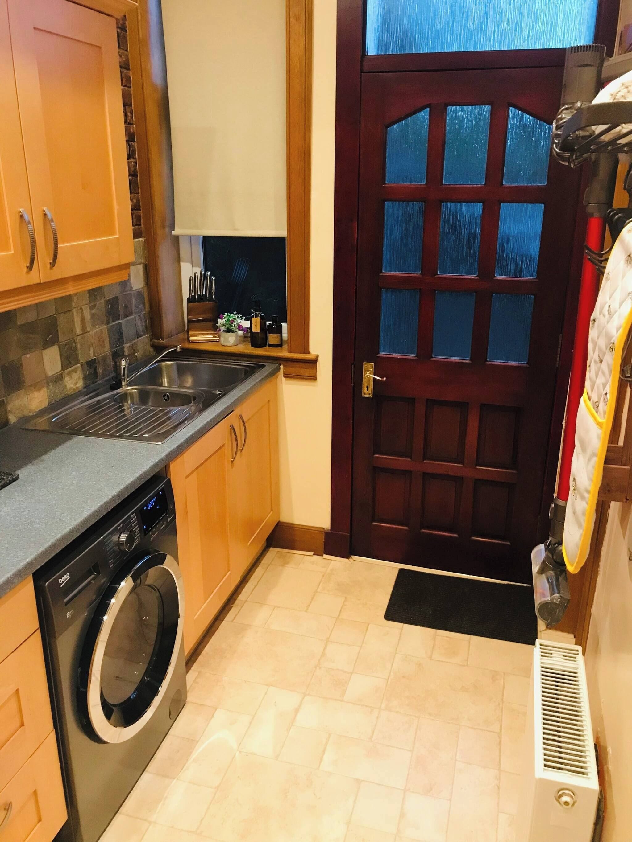 Une cuisine compacte équipée d’un lave-linge, d’un évier, de meubles en bois, d’une fenêtre et d’une porte avec des panneaux de verre.