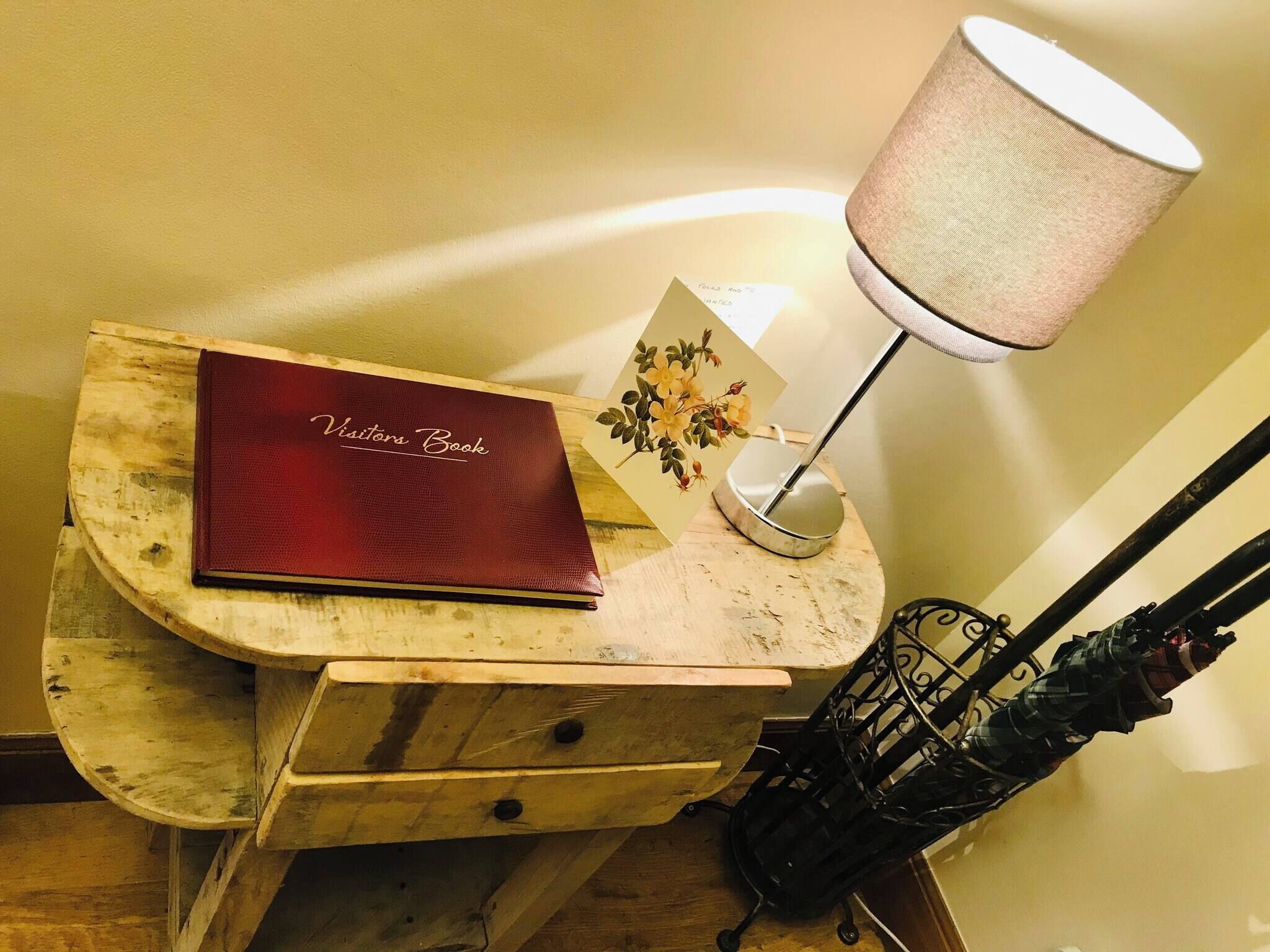 Un tavolino di legno con un libro rosso intitolato “Visitor Book” e una lampada con paralume beige.