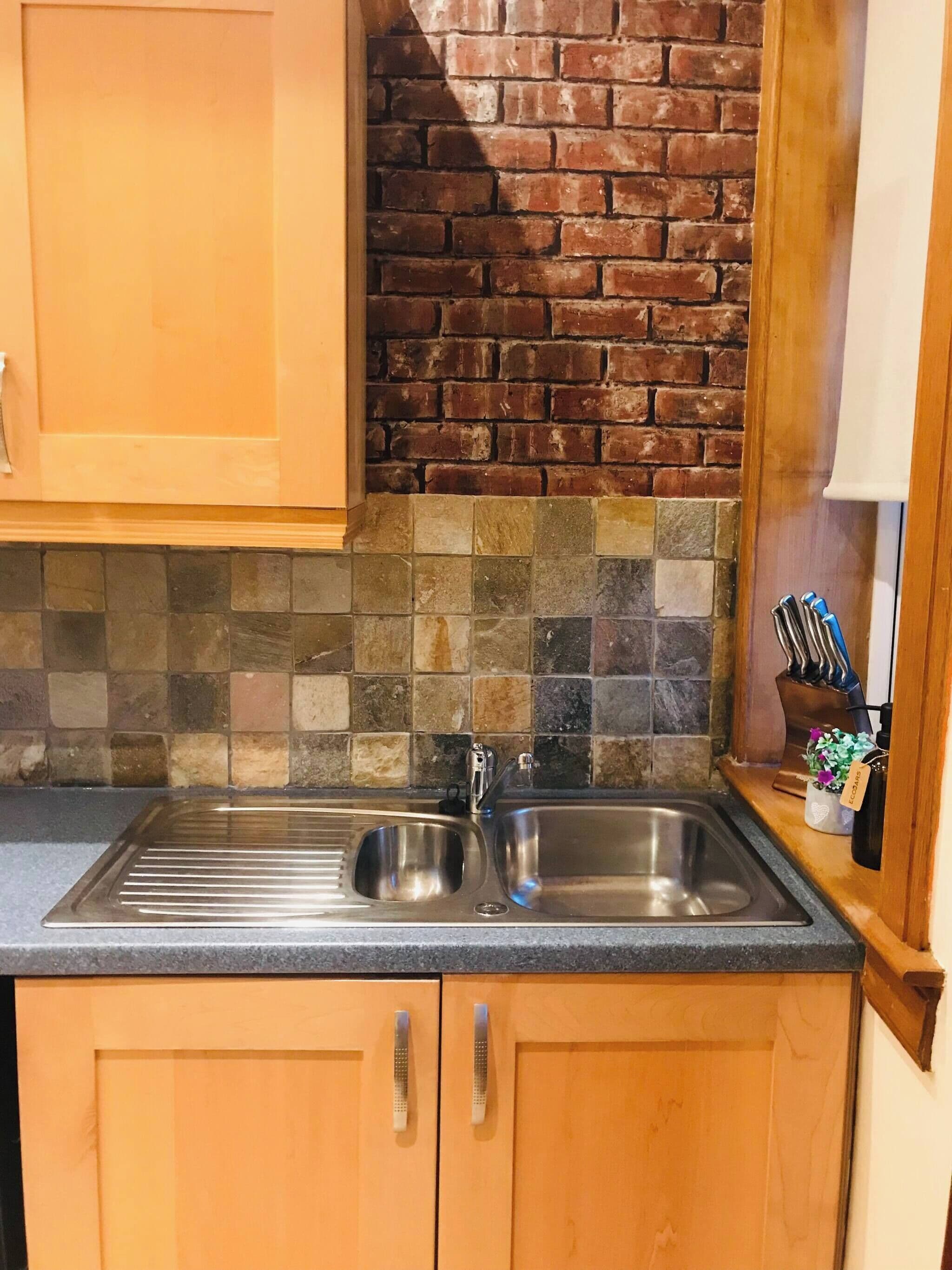 A kitchen with a stainless steel sink, wooden cabinets, a tiled backsplash, and a brick wall.