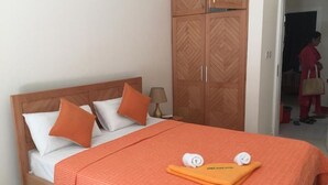 A neatly made bed with a patterned orange bedspread, two white pillows, and two orange pillows. There is a wooden wardrobe with a chevron pattern to the left of the bed. A towel and a small orange towel are placed on the bed. - West Front One No 4 With Seafront Beachfront Licensed By Tourism Authority 14977 (Flic-en-Flac)