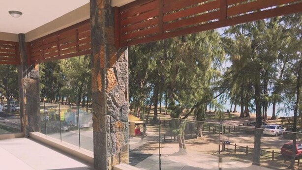 A covered outdoor area with stone pillars, glass railings, and a view of a parking lot and trees. - West Front One No 4 With Seafront Beachfront Licensed By Tourism Authority 14977 (Flic-en-Flac)