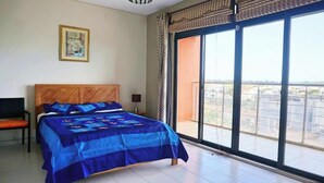 Quarto com cama grande, uma cadeira, mesa de cabeceira, quadro na parede e varanda com vista.