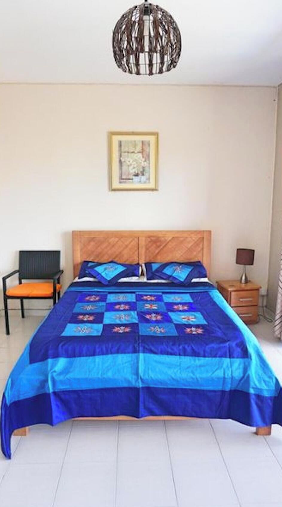 Une chambre avec un grand lit, une tête de lit en bois, un tableau encadré au mur, une chaise et une table de chevet avec une lampe.