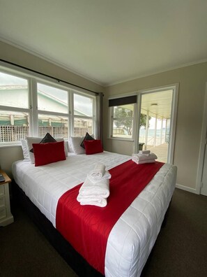 A hotel room with a large bed, two windows, a nightstand, and a view of the outdoors. - Rehutai Beach House (Paihia)