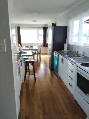 A modern kitchen with a dining area, wooden flooring, and a view of the outdoors. - Rehutai Beach House (Paihia)