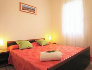 A hotel room with a bed, pillows, a nightstand, a lamp, a picture on the wall, and a window with curtains.