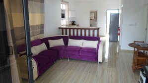 A living room with a purple sectional sofa, a wooden dining table, and a kitchen area in the background. - Residence La Colombe Vacation Rentals (Mont Choisy)