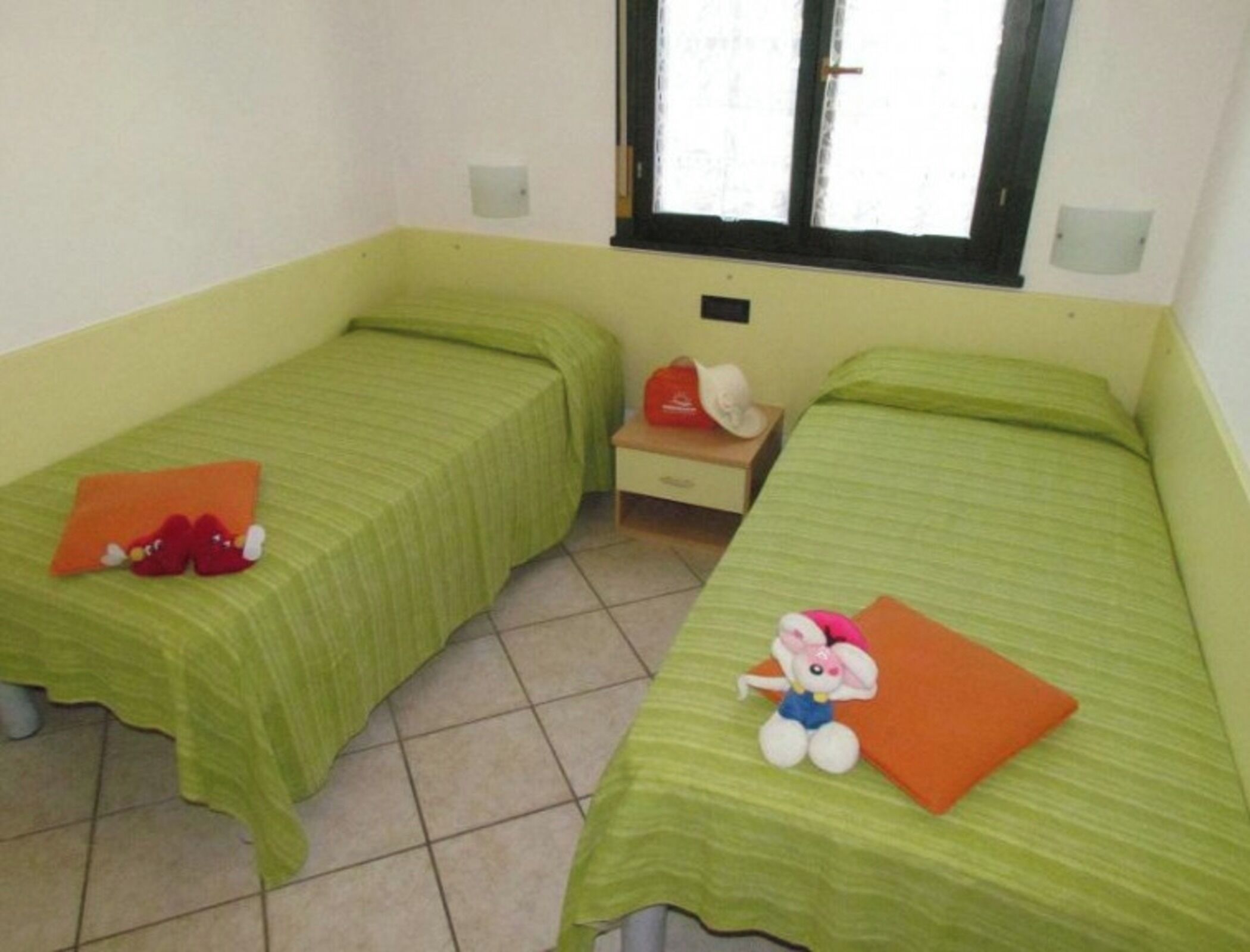 A room with two single beds, each with a green bedspread and an orange pillow. There is a small table with a red bag and a white hat on it.