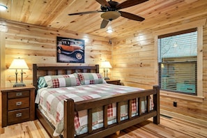 A wooden-paneled bedroom with a bed, two bedside lamps, a ceiling fan, and a window with blinds.