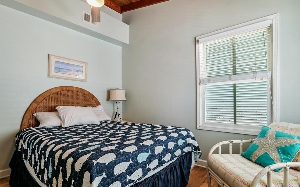 A bedroom with a bed, a wicker headboard, a bedside table with a lamp, a window with blinds, and a chair with a cushion.