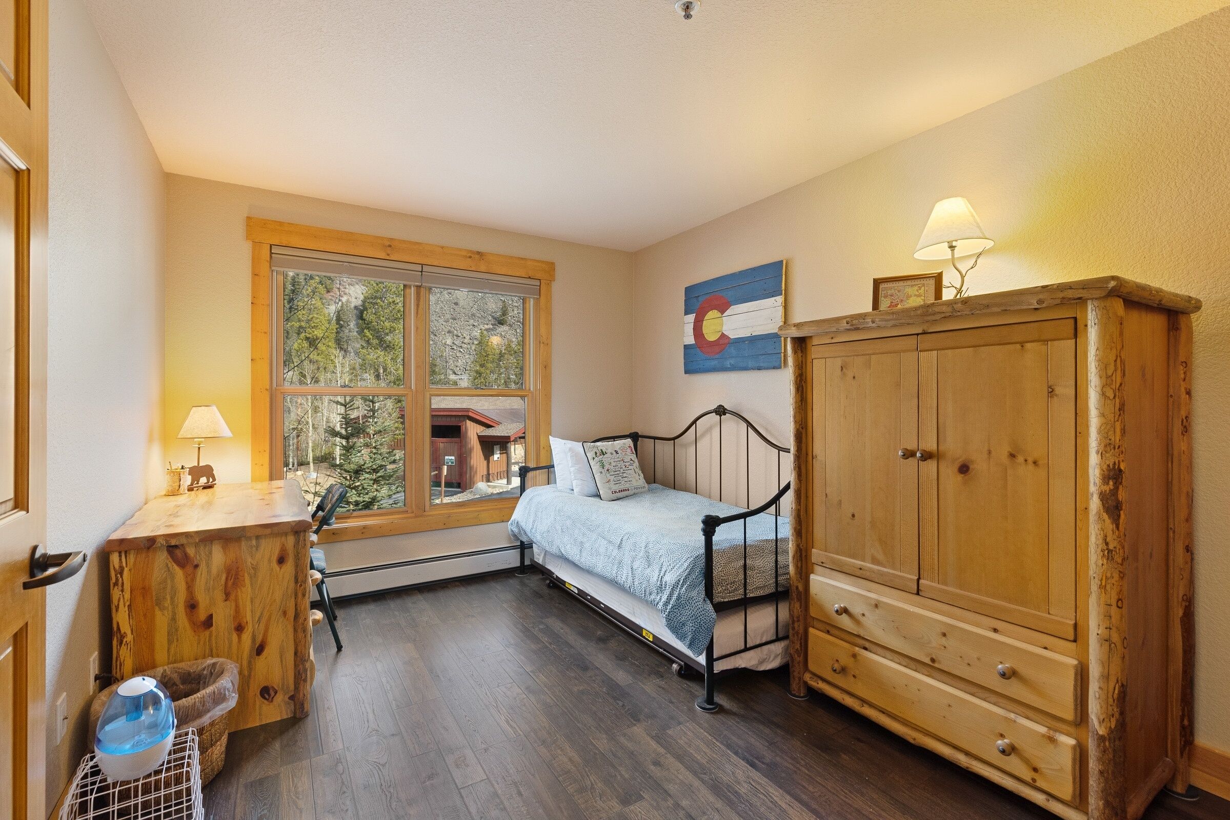 A cozy bedroom with a wooden bed, a large window, a wooden cabinet, and a small desk with a lamp.