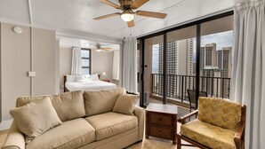 A hotel room with a large bed, a sofa, a chair, a small table, and a balcony with a city view.