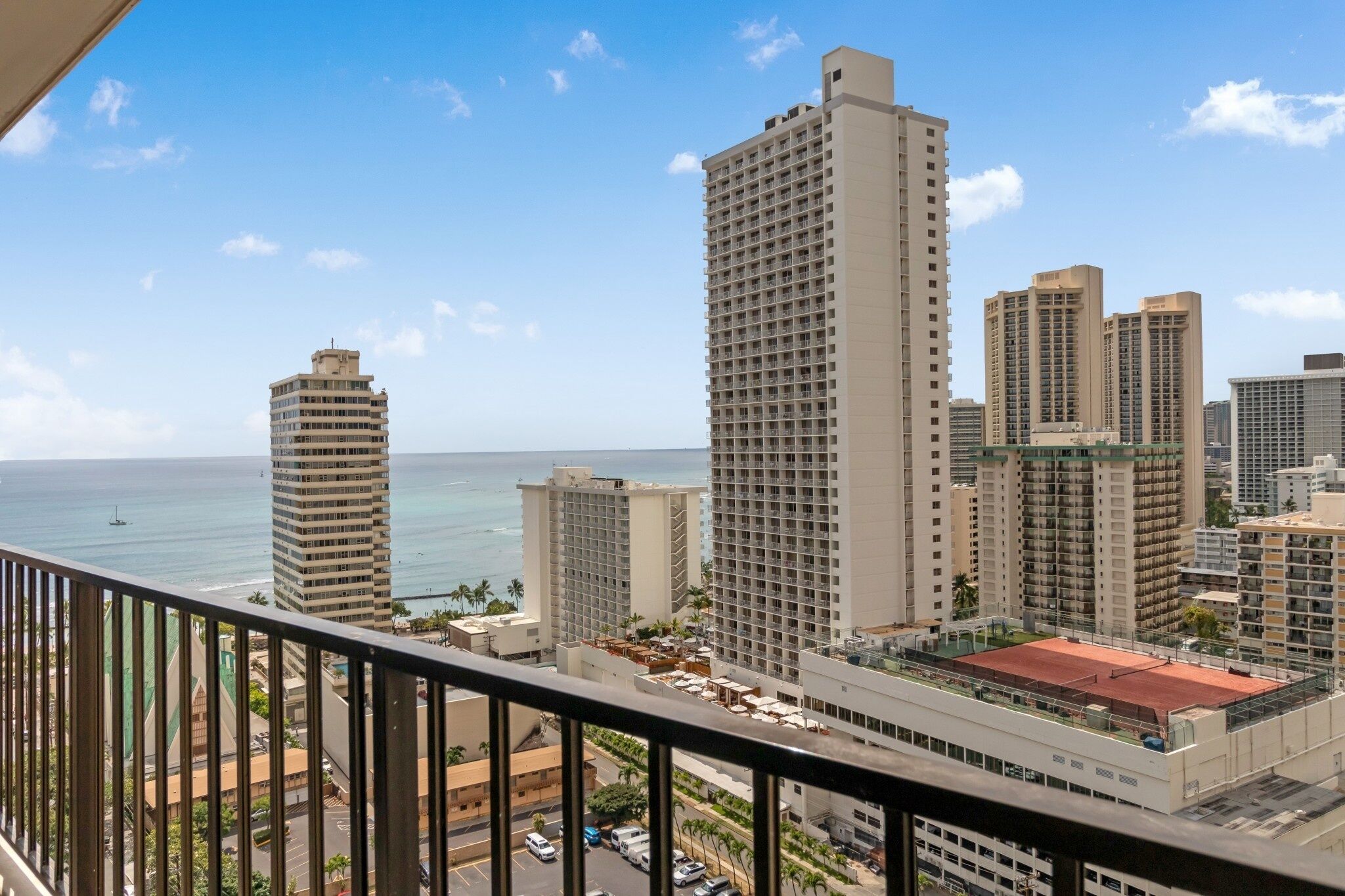 A high-rise hotel with multiple floors, a red-roofed tennis court, and a view of the ocean.
