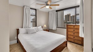 A hotel room with a bed, a wooden dresser, a ceiling fan, and large windows with curtains.