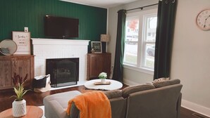 Een woonkamer met een open haard, een bank, een salontafel en een aan de muur bevestigde televisie.
