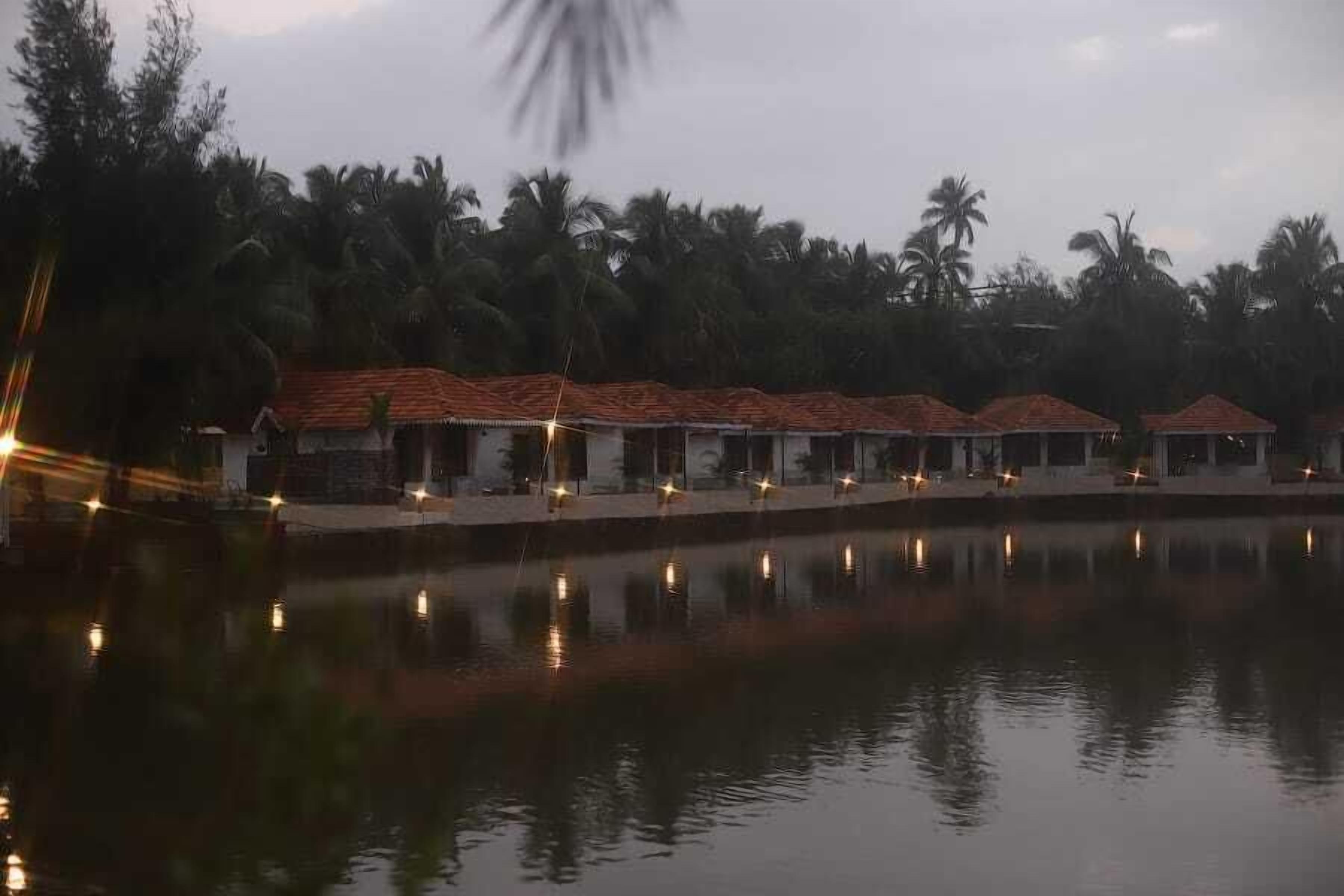 Une rangée de bâtiments de style bungalow, avec des toits recouverts de tuiles rouges, est située le long d’une étendue d’eau calme, et les bâtiments ainsi que les palmiers qui se trouvent à l’arrière se reflètent dans l’eau.