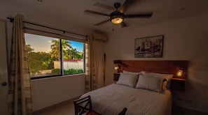 Un dormitorio con cama, una silla, ventilador de techo y ventana con cortinas.