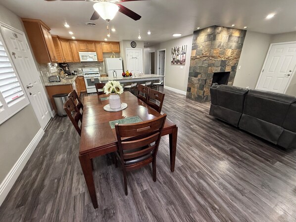 A modern kitchen with wooden cabinets, a dining table with chairs, a stone fireplace, and a gray sofa.