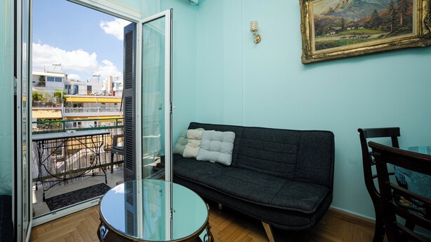 A balcony with a glass door, a dark grey sofa with cushions, a round glass table, and a wooden chair. - Acropolis view vintage style apartment (Athens)