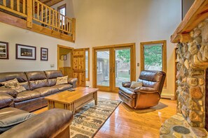 A spacious living room with a leather sofa, a leather armchair, and a wooden coffee table. There is a stone fireplace and a large window with a view of the outdoors.