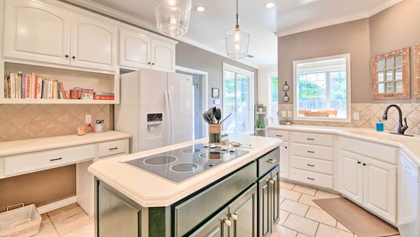 A modern kitchen with white cabinetry, a central island with a stovetop, pendant lighting, and a view of the outdoors through large windows.