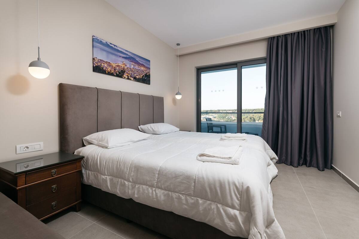 Un moderno cuarto de hotel con una cama grande, una mesita de noche, un aparador, un cuadro de un paisaje urbano y un ventanal con vistas.