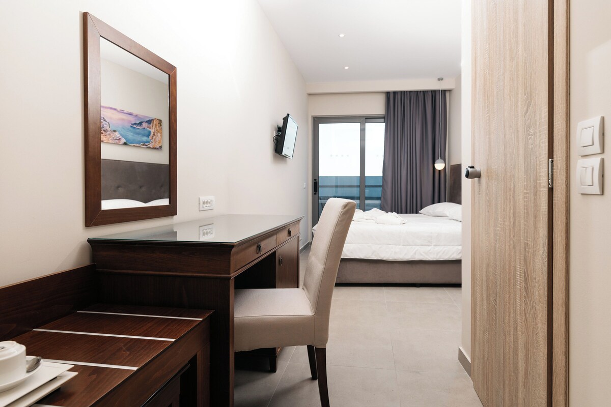 Habitació d'hotel amb escriptori de fusta, una cadira, un llit, una finestra gran amb cortines i vista al mar.