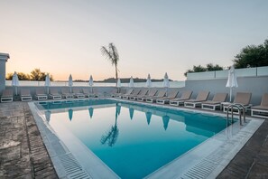 Una piscina rectangular con agua azul cristalina, rodeada de sillas de descanso y sombrillas, con vista a árboles y un cielo despejado.