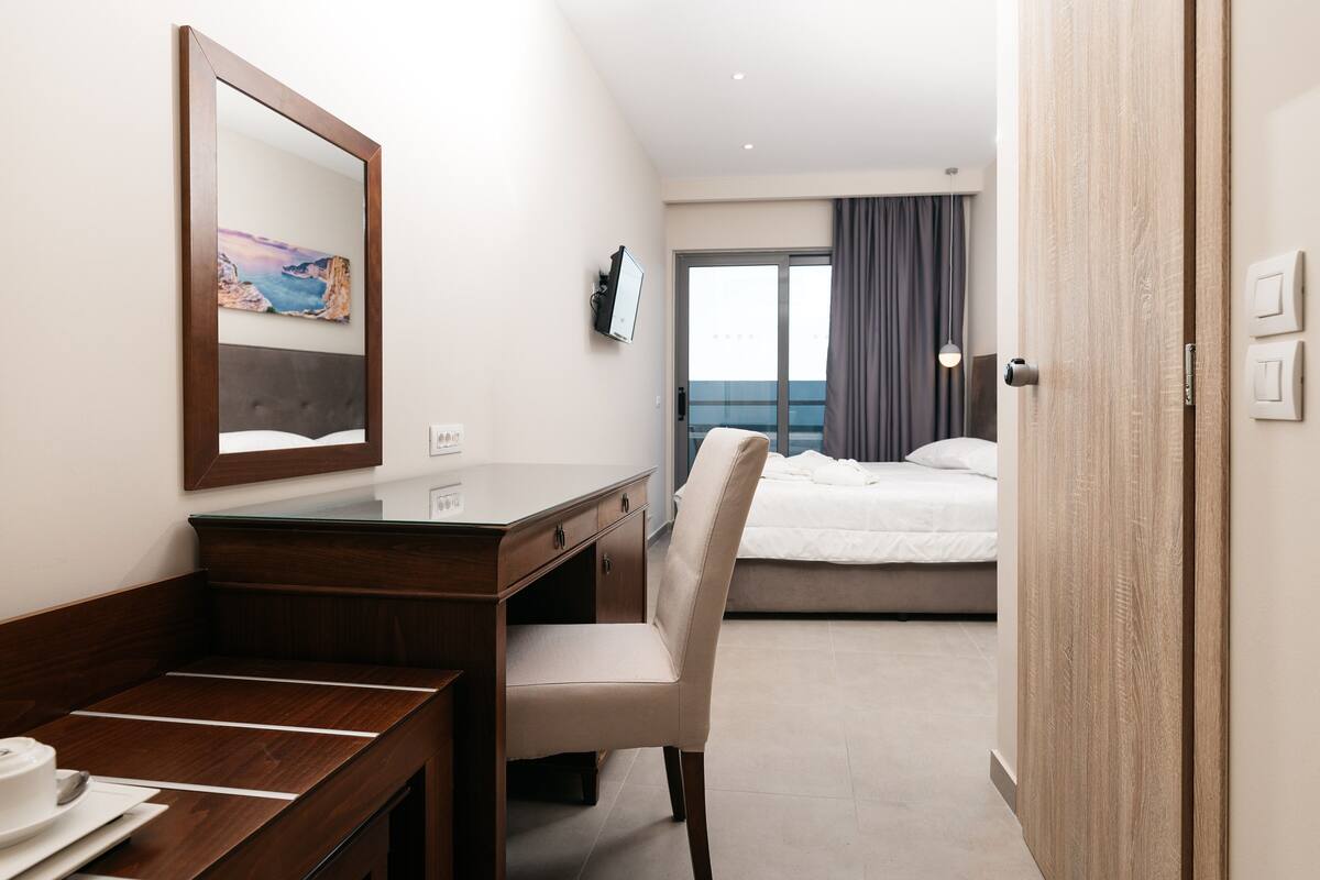 Habitación de hotel con escritorio de madera, una silla, una cama, un ventanal con cortinas y vistas al mar.