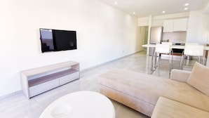 Een moderne woonkamer met een flatscreen televisie aan de muur, een bank, een salontafel en een keuken in de achtergrond.