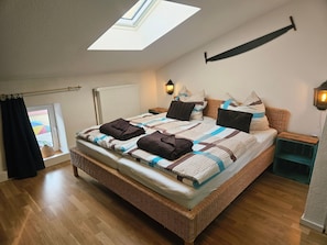 A bedroom with a skylight, a bed with a patterned bedspread, a wicker headboard, a bedside table with a lamp, and a view through a window. - Schnitterhaus Milena on Lake Flee - Milena Vacation Home on Lake Flee (Göhren-Lebbin)