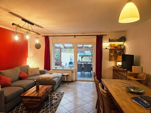A living room with a sofa, coffee table, dining table, and a TV. There is a window with curtains, a fireplace, and a bookshelf.