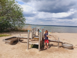 A playground with a slide and climbing structures, a sandpit, and a body of water in the background.