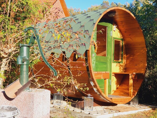 A wooden structure with a circular exterior and a green door, surrounded by trees and a metal water spigot.