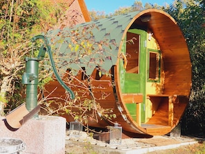 A wooden structure with a circular exterior and a green door, surrounded by trees and a metal water spigot.