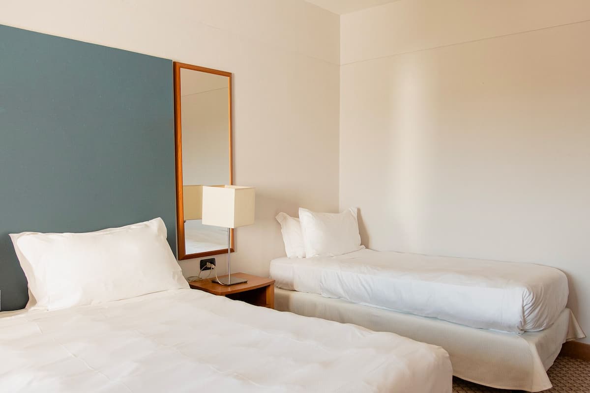 A hotel room with two single beds, a nightstand with a lamp, and a large mirror.