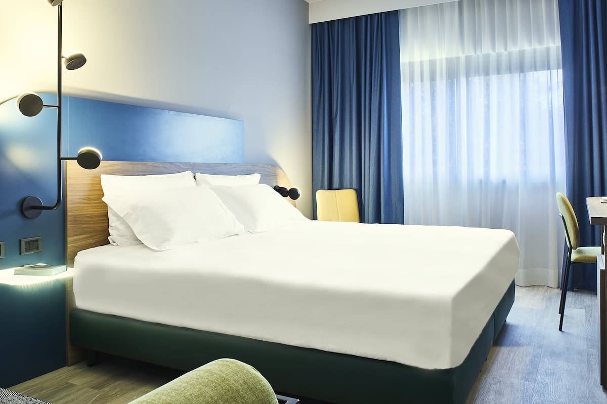 A neatly made bed with white linens, a bedside table with a lamp, and a chair.