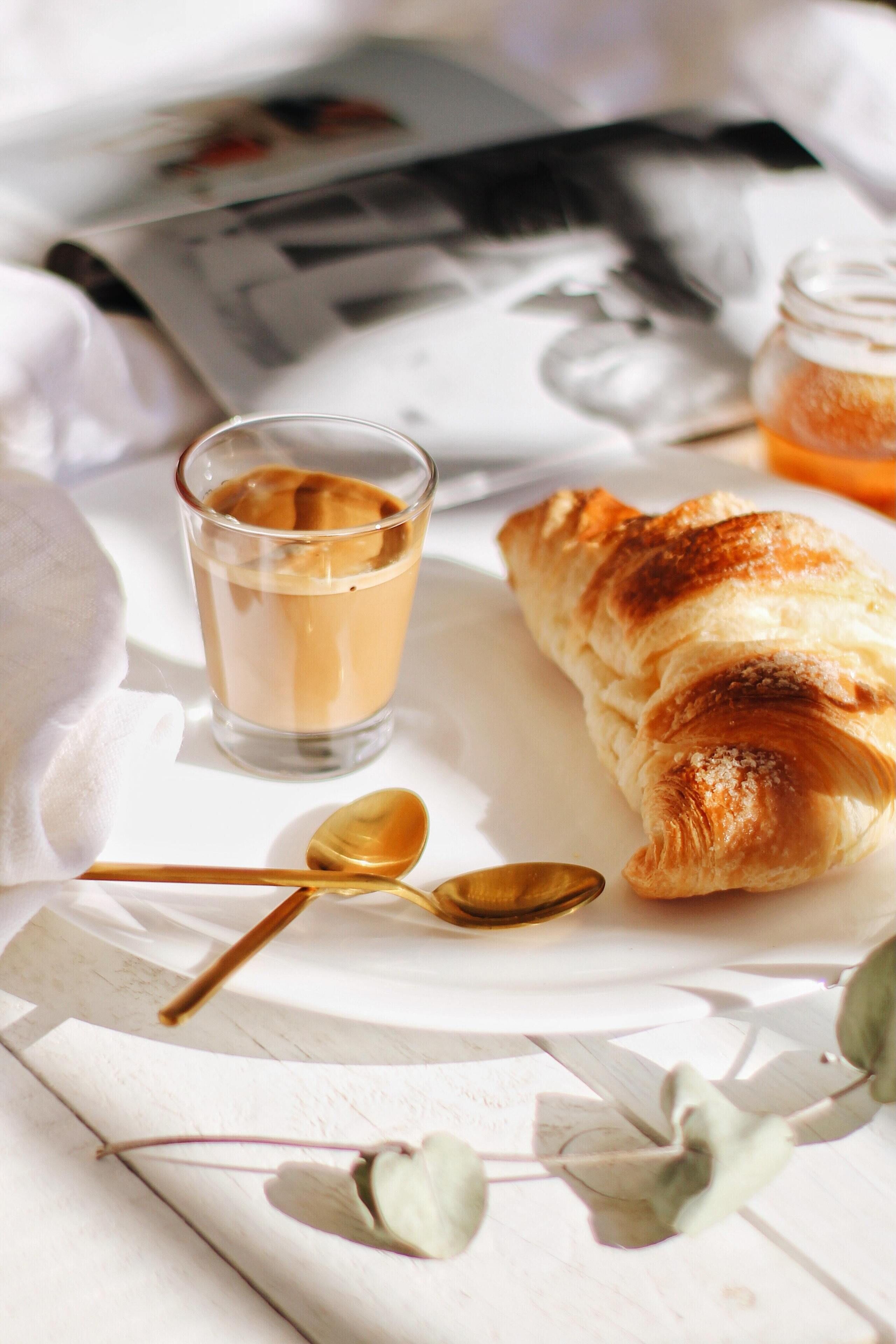 Desayuno con un croissant, una taza de café y una cuchara sobre una mesa blanca.
