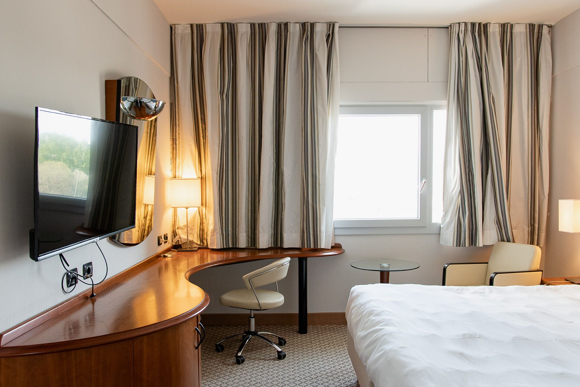 Una camera d'albergo con un letto, una scrivania con televisore, una sedia, un tavolino e tende.