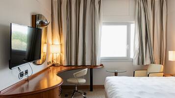 Una camera d'albergo con un letto, una scrivania con televisore, una sedia, un tavolino e tende.