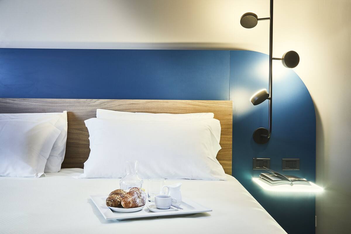 A neatly made bed with white linens, a tray with pastries and a pitcher of coffee, and a wall-mounted light fixture.
