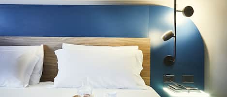 Cama arrumada com lençóis brancos, bandeja com doces e um jarro de café, e luminária fixada na parede.