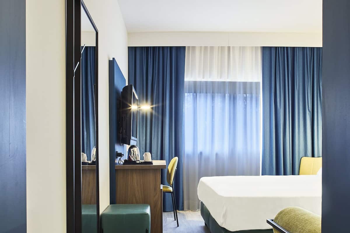 A hotel room with a bed, a desk, a chair, a mirror, and a window with curtains.