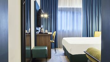 Una habitación de hotel con cama, escritorio, silla, espejo y ventana con cortinas.